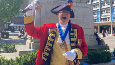 A town crier in traditional costume in a town square ringing a bell in a square with a stone plinth behind him.