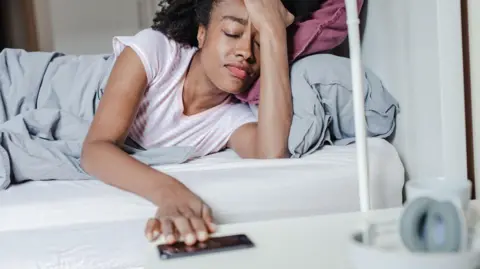 A stock image of a woman waking up in bed and reaching for a smartphone