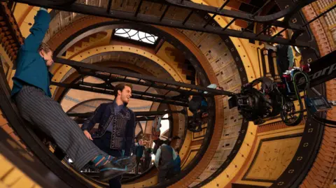 Actors inside a circular stage built to look like a library, with a film camera to the right of the picture