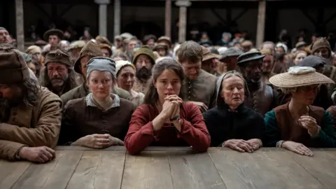 Universal Studios A film still from Hamnet, showing Jessie Buckley as Agnes (Anne Hathaway). The picture shows a group of characters who are audience members, pressed against the stage of the Globe. Agnes is in the middle in a red dress with her hands pressed together, looking sombre.