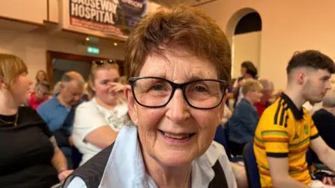 Carol Wallace has short brown hair and black glasses, she is wearing a grey pinstripe waistcoat over a light blue blouse. She is sitting in a busy meeting in Coleraine Town Hall.