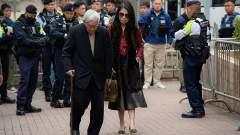 Getty Images Jimmy Lai's wife Teresa (right) and Joseph Zen, cardinal of the Holy Roman Church, arrives at court on 9 Feb, Monday, for Lai's sentencing hearing. Teresa Lai is wearing black sunglasses, a pink shirt and black skirt, while Zen is in a black suit, holding a cane.
