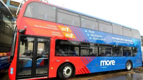 Morebus A side view of a large double decker local bus in Bournemouth. It is painted blue and red, and has a handful of destinations like Poole and Westbourne printed on the side. 