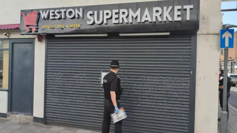 A police officer wearing a black uniform, hat and blue latex gloves. He is standing outside the Weston Supermarket, attaching a closure order to the black corrugated shutters.