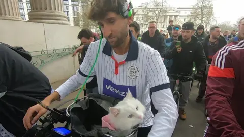 A close-up of a man wearing headphones and cycling through London with DJ decks on the front of his bike and a white cat in a basket, looking out to the side towards the camera. Other people are riding behind and alongside them as part of an event