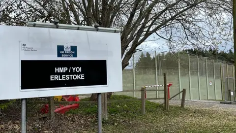 Karen Gardner/BBC The exterior of HMP Erlestoke with a large sign outside and high barbed-wire fences. There is a tree behind the white and black sign, which says HM PRISON SERVICE and HMP / YOI ERLESTOKE.