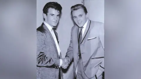 Ricky Ford A young Ricky, right, wearing a 1950s suit and tie. He is shaking hands with Billy Fury, right, who is also wearing a suit and tie, and has his hair styled in a 1950s quiff