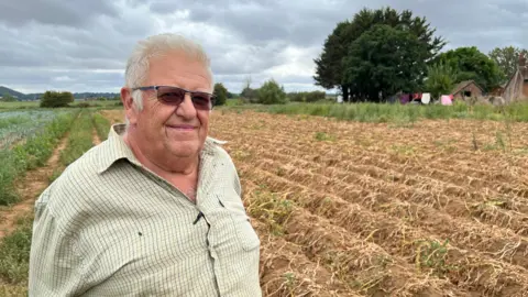 An older man wearing a light shirt sands in front of a brown field where the crops have failed.