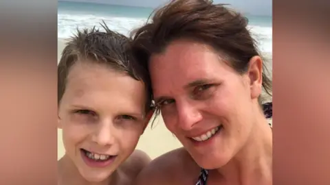 A mum and her young teenage son smile for a selfie as they stand on a beach abroad with the ocean behind them