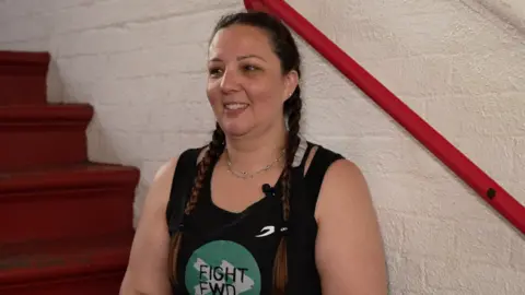 Saria Alireza sits on a red staircase inside a boxing gym, wearing a black Fight Forward vest and yellow hand wraps, smiling during an interview.