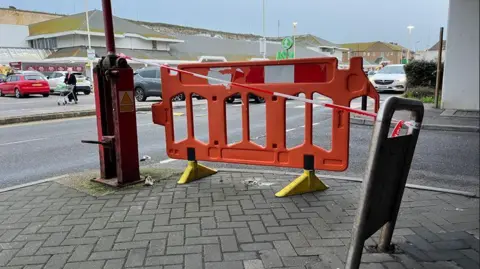 A red plastic barrier in place at the edge of a car park with shoppers and cars in the background. 