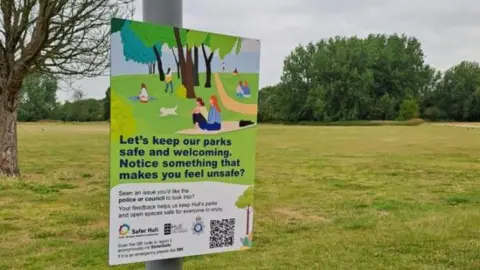 One of the signs on a grey post in a park area. The sign has a drawing of people enjoying the park and says "Let's keep our parks safe and welcoming. Notice something makes you feel unsafe?" The QR code is below the writing.