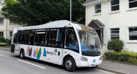 BBC Bus parked near the bus stop near the house. The bus and the house behind it are painted white.