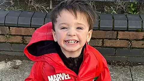 Jude Gerrard, a young boy with brown hair, grins as he is being photographed. He is wearing a red waterproof coat with Marvel branding.