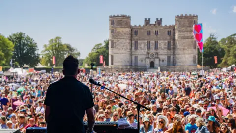 A performer is on stage at a keyboard and microphone with his backturned. Beyond him is an enormous crowd of people. Behind the crowd there is a large castle. 