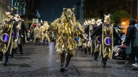 DCSDC Shows more than a dozen people dressed as wolves in golden and black outfits walking through a street. 