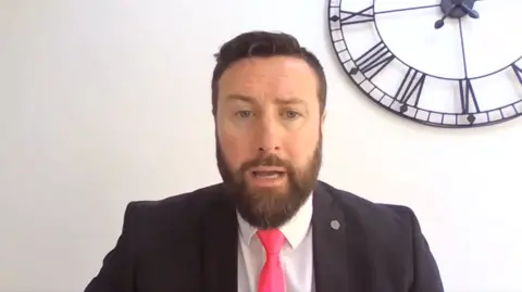Damian Kenny, 45, who has dark brown hair swept to one side and a dark brown beard, and is wearing a black suit jacket, white shirt and pink tie, speaks into a camera with a plain white wall and a large black clock in the background. 