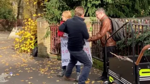 The protester, a woman with blonde hair and a red puffer jacket and jeans, holding a large placard referecing the war in Gaza, two men are trying to take the placard off her