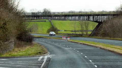 Underbarrow Road Bridge over the A591. A single car drives on the other side of the road. There is a short patch of grass in between the double carriageway. There is a small junction to the left.