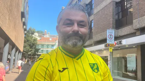 Maddy Jennings/BBC Simon Badderly smiles directly at the camera, wearing a yellow Norwich City football shirt. He is standing in a street. 
