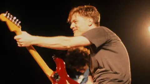 Getty Images Bryan Adams performs on stage while playing an electric guitar. He is wearing a brown T-shirt and is looking away from the camera.