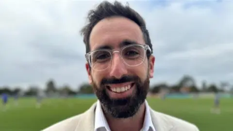 Zac Seidler grins in a close-up. He has a beard and moustache and wears clear glasses, a cream jacket and white shirt with an orange moustache pin attacked to the jacket. 