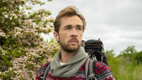 Thomas Winward A close-up shot of James, a young bearded gentleman, standing in a field full of green shrubbery and pink flowers. He is wearing a fleece with red, blues and grey on it, and is carrying a rucksack.