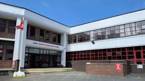 BBC The entrance to the UCM building, which is build of brown bricks and features while concrete cladding and pillars, The entrance is glass-fronted and has UCM University College Isle of Man written in black and red above it.