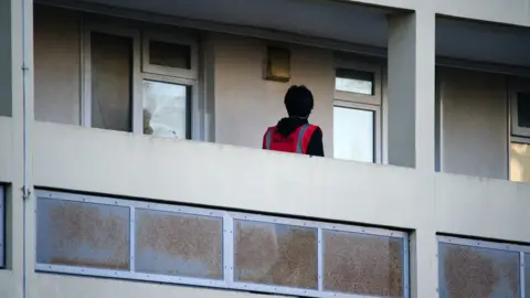 PA Media A man walking along a balcony at Barton House in Bristol after it was evacuated in 2023. He has dark hair and is wearing a pink high vis vest. 