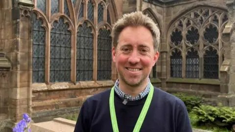 A man wears a checked shirt underneath a navy jumper with a yellow lanyard round his neck. He has short blond hair and is standing in front of a cathedral building with ornate leaded windows and stone walls.