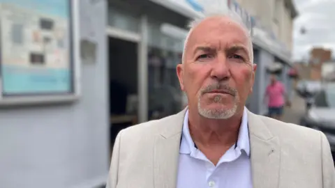 This image shows Julian Wheeler, a man with white hair and a grey goatee beard, standing outside his shop wearing a white polo shirt and cream-coloured jacket.