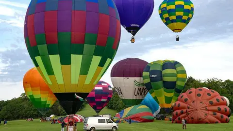 Claire Hartley A number of colourful hot-air balloons in the air and on the ground in the middle of a field.