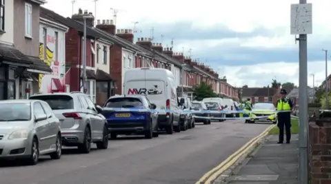 BBC A road with a row of terraced houses along it in Gorse Hill. There is a police cordon across the road and a police officer standing with a police car behind them in the distance. 