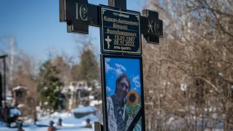 Matthew Goddard/BBC Vitaly died fighting against the Russians in eastern Ukraine. The photo shows his smiling photo attached to a crucifix marking his grave in a snowy cemetery in Kyiv
