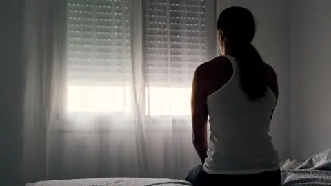 Alvaro Medina Jurado A woman with long brown hair in a ponytail is sat on the edge of the bed facing a white wall and window.