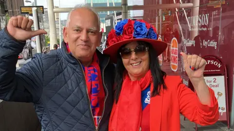 Eugene Borges in a quilted blue jacket and a club scarf. Pamela Gould stands next to him in a red hat covered in red and blue roses. She wears a club t-shirt, red blazer and scarf. 