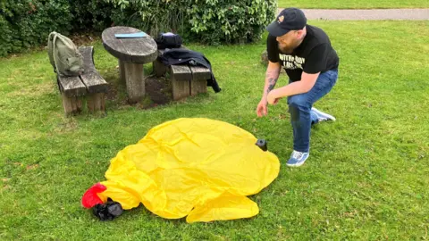 Liam Birkett, wearing dark jeans, a black tshirt and baseball cap and has a brown beard, about to inflate the duck on a patch of grass