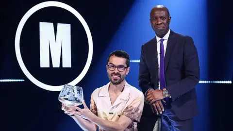 Two men are in a room, with one sitting down holding a decorative 
glass bowl. He has short hair and is wearing a patterned shirt and glasses. The second man is standing behind him and wearing a dark suit, white shirt and purple tie. A large "M" is in a white circle on the wall in the background.