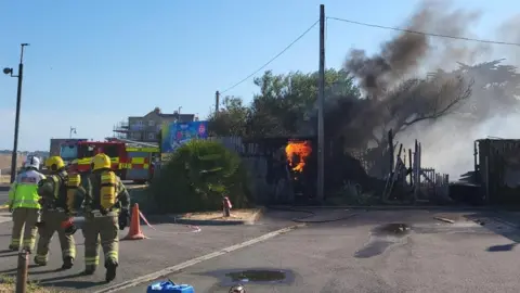 DWFRS Firefighters wearing breathing apparatus walk towards a fire engine as flames and smoke rise from an electricity substation.