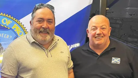 A man with dark hair and dark facial hair wearing all grey, standing next to a smaller bald man who is wearing navy with a saltire on his polo shirt, they are standing in front of a saltire with the world providence written on it. 