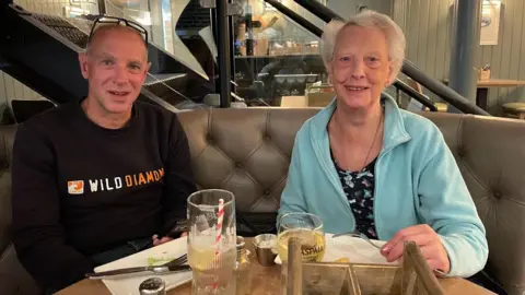 Denise Baxter Ewan Baxter and his mum Margo sit at a table in a restaurant. Drinks abnd plats are in front of them. He is a bad man, with glasses resting on top of his head and wearing a black top with white and orange lettering. Margo is a woman with white hair, wearing a patterned dress and light blue/aqua jacket. 