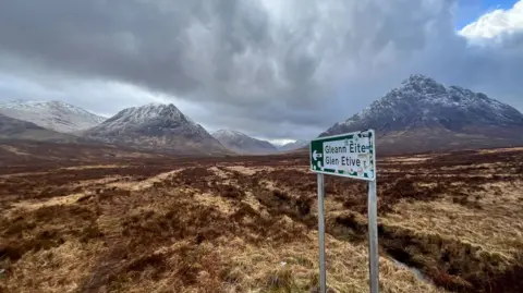Michael Thomson A wide moorland valley beneath dark, heavy clouds, surrounded by snow‑dusted mountains. In the foreground stands a weathered road sign pointing left to “Gleann Èite / Glen Etive”.