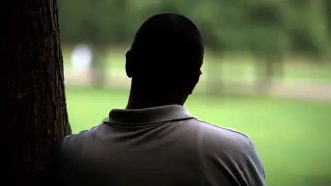 The silhouette of a man standing, leaning against a tree and looking out at a blurred park
