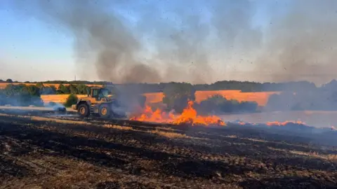 Large fire on a farm, there is a yellow tractor and lots of smoke in the photo