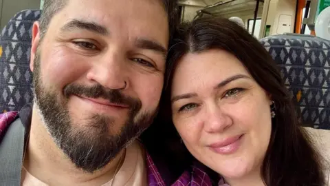 Chris Sherwin, who has beard and black hair, and Amy Hawkswood, who has long dark hair, sitting together on a train carriage, smiling. The pattern of the seat fabric is behind them.