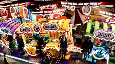 A carnival cart with dancers standing on it. The carts are purple, yellow and green and have lights attached to them.