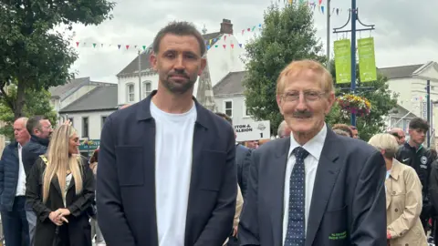 Two men pose for a photo. One is wearing a white top and a navy jacket and one is wearing a suit. Crowds are behind them.