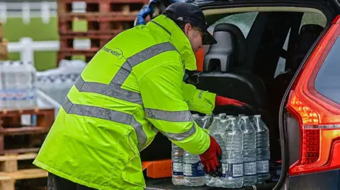 Getty Images Man wearing yellow hi-viz jacket with South East Water logo on the back loads bottled water into the boot of a car