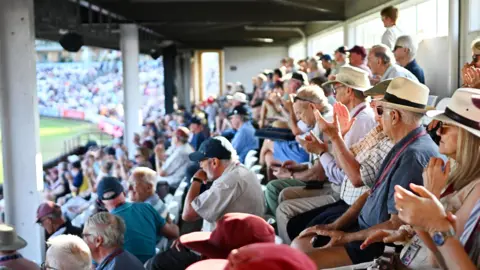 Getty Images Dozens of Somerset cricket fans sit in a stand at Taunton on a sunny evening as they watch their team play Gloucestershire. Many of them are wearing hats and sunglasses