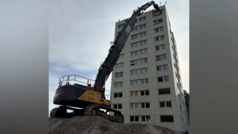 Hull City Council A digger is pointed at the top of a high block of flats, about to knock it down. It is positioned on a high mound of soil.